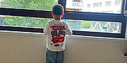 Ibrahima participe au concours pour gagner de l'argent avec cette photo : child, boy, standing, window, indoor, shirt, jeans, sneakers, floor, wood_floor, building, tree, apartment, daylight, back_view, casual_clothing, young_child, real_photo, urban, curtain