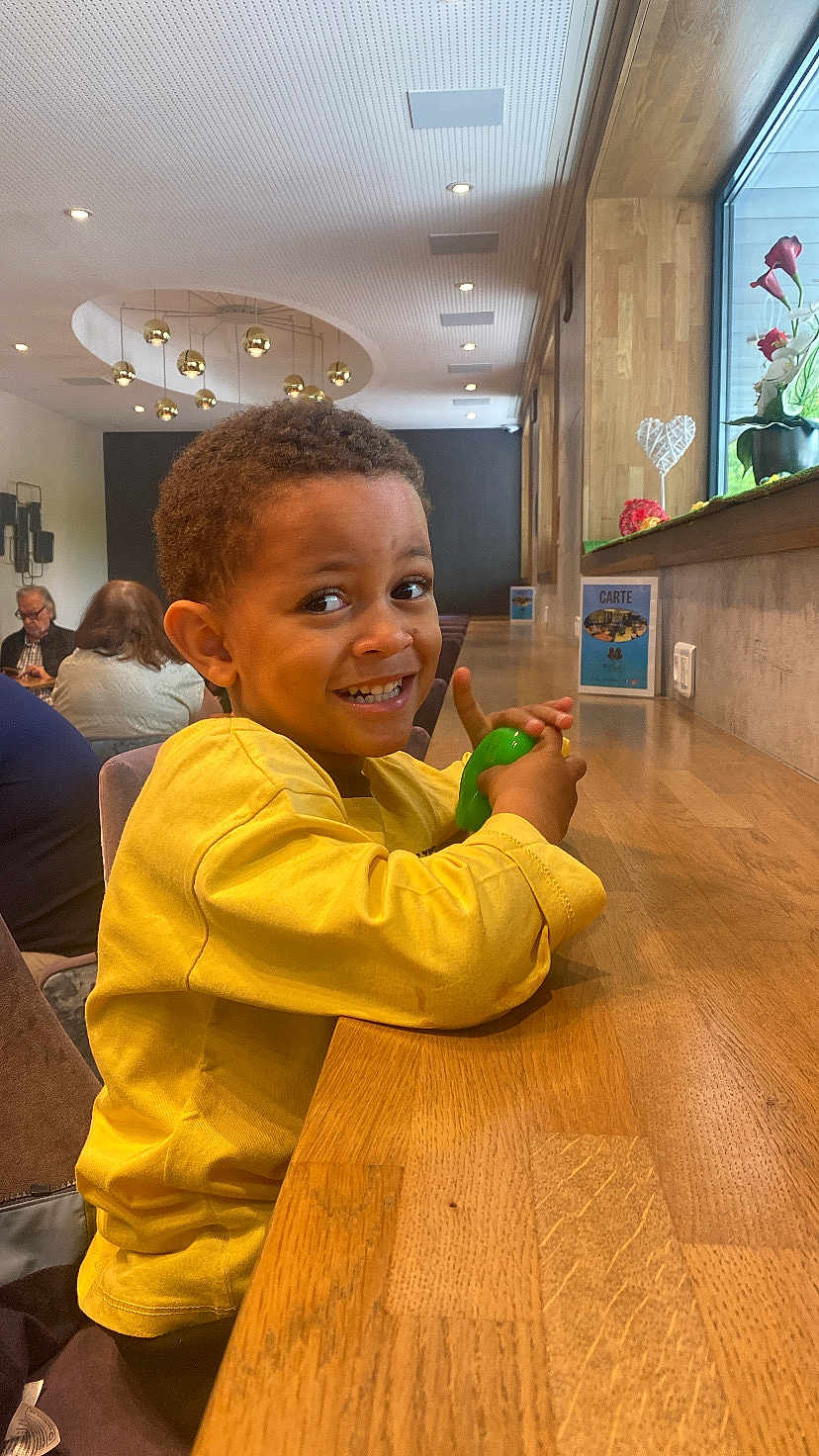 Ibrahima participe au concours pour gagner de l'argent avec cette photo : cafe, casual, child, decor, face, happy, indoor, lighting, modern, people, person, playful, portrait, restaurant, seated, smiling, toy, window, wooden_counter, yellow_shirt