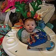 Emerald is registered to the contest to win money with this photo: baby, infant, reindeer_antlers, walker, toy, blanket, living_room, child, smile, face, headwear, holiday, gift, plush_toy, seated, clothing, person, indoor, cute, happy