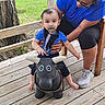 Miguel is registered to the contest to win money with this photo: baby, pacifier, toy_bull, wooden_deck, adult, outdoor, greenery, wooden_railing, blue_shirt, glasses, shorts, sneakers, child, playful, supportive, crouching, cute, smiling, person, daylight