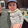 baby, child, hat, onesie, person, indoor, kitchen, cabinet, adult, holding, face, expression, cute, clothing, hand, toy, countertop, appliance, smile, portrait