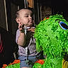 toddler, child, piñata, green, bird, toy, indoor, night, window, curious, clothing, shorts, patterned_shirt, face, hand, expression, colorful, party, celebration, playful