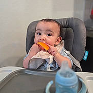 Miguel is registered to the contest to win money with this photo: baby, child, high_chair, bib, orange, food, eating, sippy_cup, table, indoor, curious, cute, messy, mealtime, infant, person, face, hands, feeding, kitchen