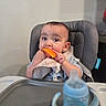 baby, child, high_chair, bib, orange, food, eating, sippy_cup, table, indoor, curious, cute, messy, mealtime, infant, person, face, hands, feeding, kitchen
