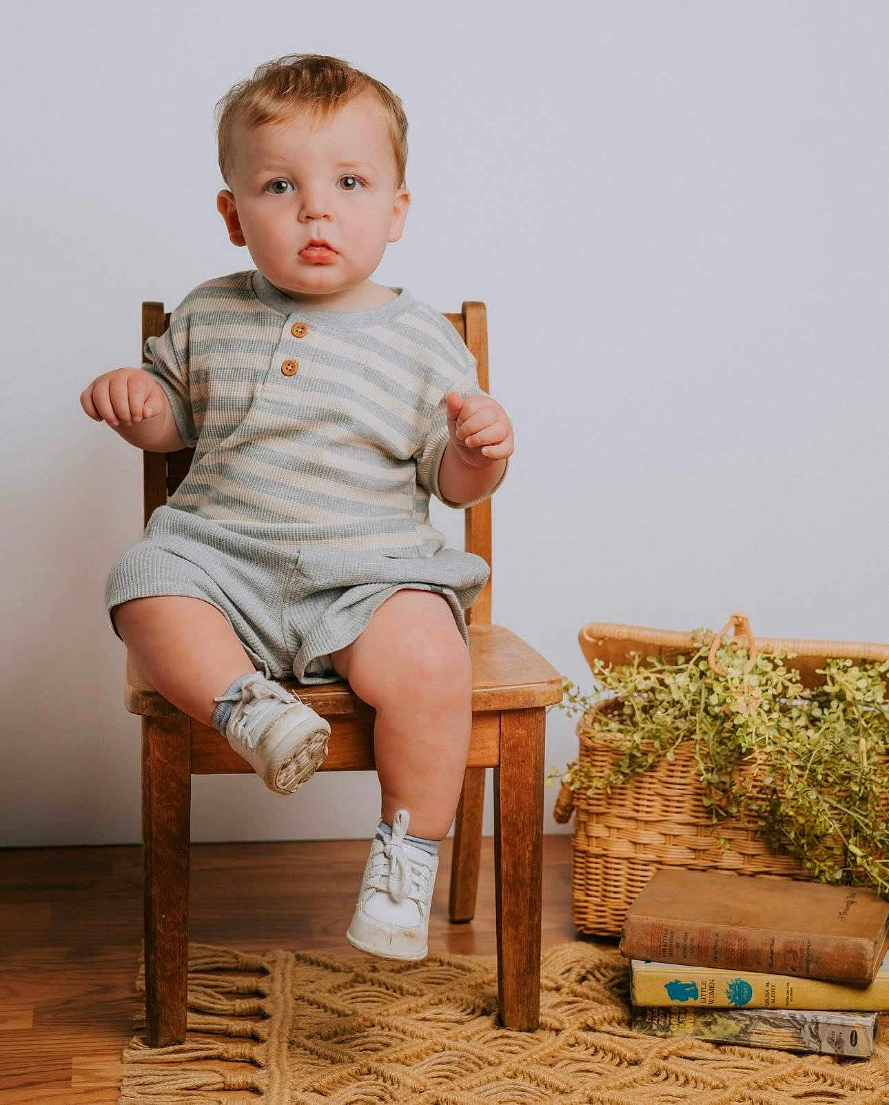 Noah joined the competition — help win amazing prizes! toddler, child, chair, wooden_chair, striped_shirt, shorts, white_sneakers, basket, greenery, plants, books, vintage_books, rug, floor, indoor, portrait, curious_expression, seated, baby, cozy