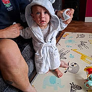 Sebastian joined the competition — help win amazing prizes! baby, child, robe, hood, bear_ears, play_mat, animal_pattern, adult_hand, couch, indoor, floor, foot, person, clothing, supporting_hand, wood_floor, toy, colorful, expression, cute