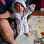 baby, child, robe, hood, bear_ears, play_mat, animal_pattern, adult_hand, couch, indoor, floor, foot, person, clothing, supporting_hand, wood_floor, toy, colorful, expression, cute
