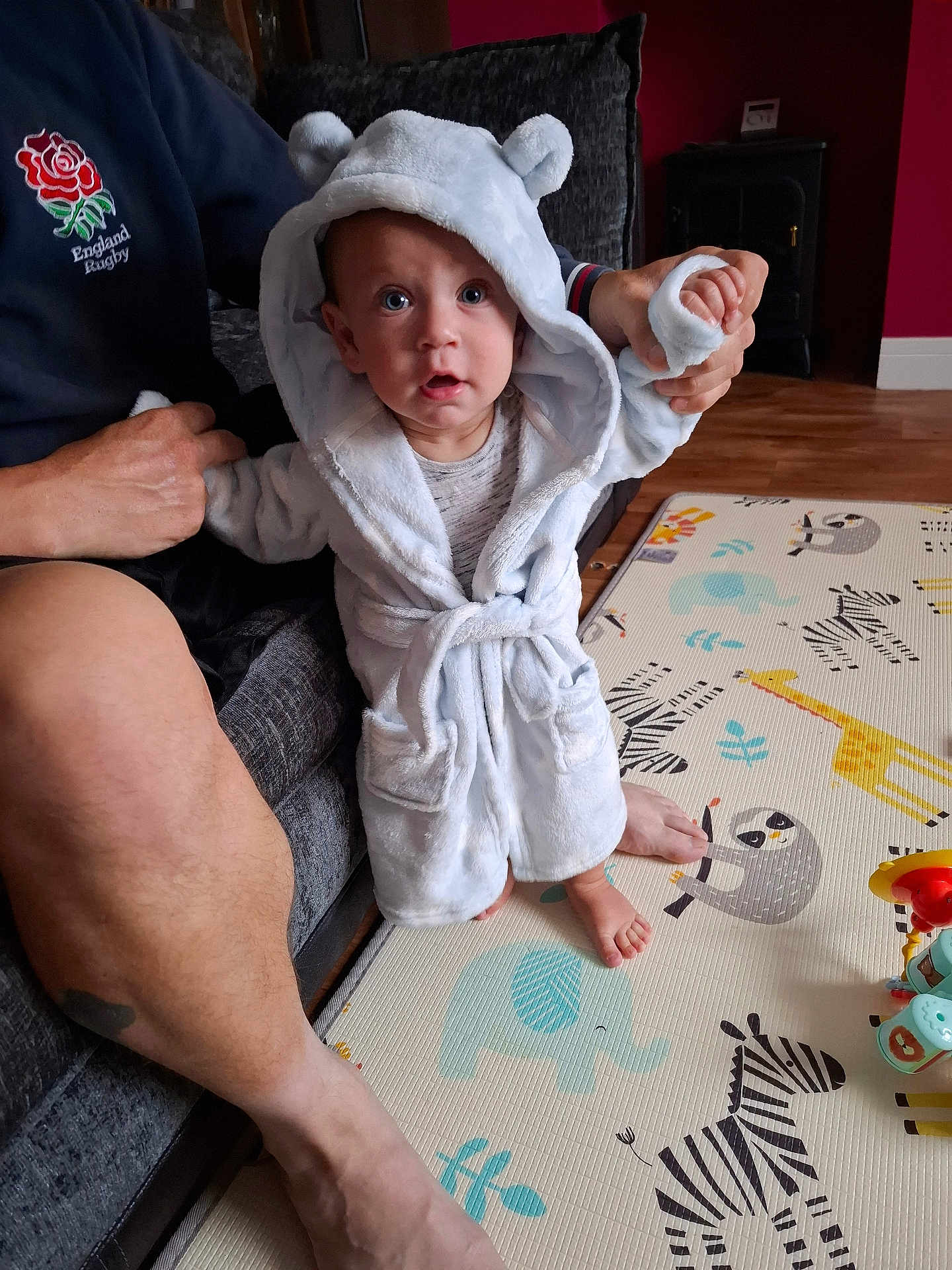 Sebastian joined the competition — help win amazing prizes! baby, child, robe, hood, bear_ears, play_mat, animal_pattern, adult_hand, couch, indoor, floor, foot, person, clothing, supporting_hand, wood_floor, toy, colorful, expression, cute