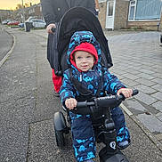Sebastian is registered to the contest to win money with this photo: adult, child, daylight, dinosaur_pattern, footwear, hood, jacket, outdoor, pavement, person, residential_area, sidewalk, smile, snowsuit, street, stroller, suburban, toddler, tricycle, winter_clothing