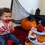 birthday_candle, cake, celebration, child, cupcakes, curly_hair, decorations, eyeballs, festive, fun, halloween, indoor, jeans, messy, pumpkins, red_frosting, smiling, spider, toddler, white_tank_top