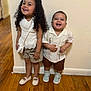child, children, smiling, happy, indoor, wall, floor, wood_floor, curly_hair, shorts, shirt, shoes, sneakers, bow, purse, siblings, standing, casual_clothing, portrait, cute