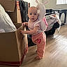 baby, child, pink_clothing, snail_backpack, cardboard_box, wooden_floor, indoor, window_light, playpen, blanket, toy, barefoot, curious, standing, home, infant, cute, decor, person, young_child