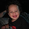 child, toddler, smile, curly_hair, teeth, black_tshirt, blanket, indoor, portrait, happy, face, person, baby, cute, infant, dark_background, cozy, relaxed, skin, eyebrows