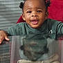toddler, child, smiling, indoor, glass_table, reflection, hair_buns, green_sweater, beige_pants, red_chair, happy, cute, portrait, person, furniture, window_blinds, skin, face, hands, seated