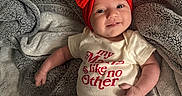 Vaeda is registered to the contest to win money with this photo: baby, infant, smiling, red_bow, headband, onesie, text_on_clothing, blanket, gray_blanket, soft_texture, cute, happy, lying_down, human, skin, small_hands, small_feet, sock, indoors, portrait