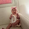 baby, child, smile, pink_headband, letter_d, armchair, knitted_blanket, soft_texture, indoor, happy, infant, cute, light_clothing, cozy, seated, furniture, portrait, person, home, relaxed