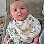 baby, infant, bib, koala, animal_print, couch, blanket, clothing, face, person, indoor, child, cute, sitting, home, soft_fabric, expression, skin, legs, hand