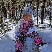 Meilani is registered to the contest to win money with this photo: toddler, child, snow, winter_clothing, mittens, hat, forest, trees, outdoor, rock, smiling, sitting, pants, shoes, nature, daylight, cold_weather, person, playful, cute
