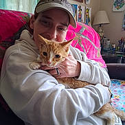 Luca is registered to the contest to win money with this photo: person, cat, indoor, couch, hugging, cap, hoodie, orange_cat, white_cat, artwork, lamp, blanket, wall, hand, watch, ring, cozy, home, furniture, smiling