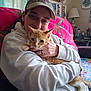 person, cat, indoor, couch, hugging, cap, hoodie, orange_cat, white_cat, artwork, lamp, blanket, wall, hand, watch, ring, cozy, home, furniture, smiling