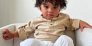 Niko is registered to the contest to win money with this photo: toddler, child, curly_hair, beige_shirt, white_pants, armchair, furniture, indoor, portrait, person, cute, baby, relaxed, seated, looking_at_camera, expression, skin, foot, hand, cozy
