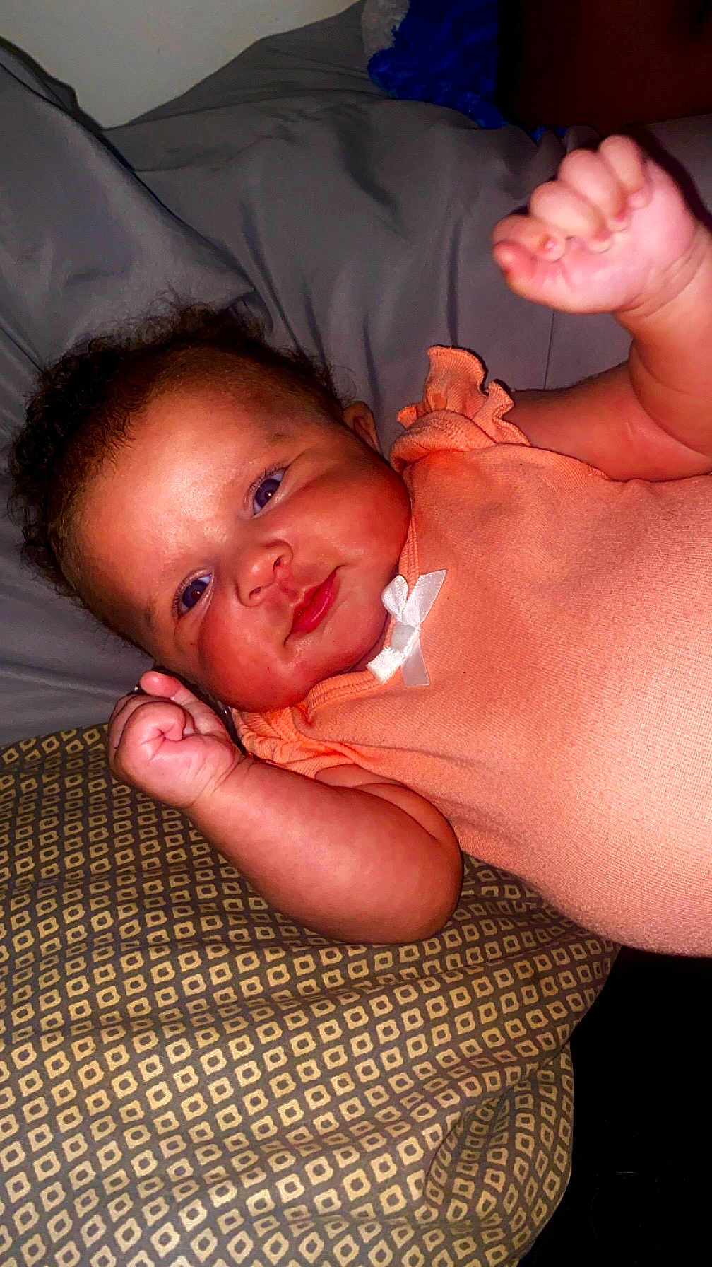 Aaliyah joined the competition — help win amazing prizes! baby, infant, child, face, fist, clothing, peach_outfit, pillow, pattern, blanket, skin, head, arm, person, indoor, cute, smile, lying_down, newborn, raised_fists
