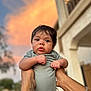 Josiah is registered to the contest to win money with this photo: baby, infant, child, person, arms, tattoo, bracelet, onesie, outdoor, sunset, sky, clouds, building, blurred_background, portrait, expression, cute, holding, human, skin