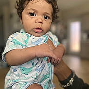 Josiah joined the competition — help win amazing prizes! baby, infant, curly_hair, onesie, whale_pattern, hand, bracelet, indoor, blurred_background, portrait, child, cute, skin, face, eyes, mouth, person, holding, young, closeup