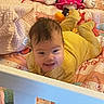 Elliana is registered to the contest to win money with this photo: baby, child, crib, bed, blanket, stuffed_toy, smile, yellow_clothing, indoor, happy, face, person, toy, plush, head, hair, cushion, soft, toddler, colorful