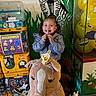 child, girl, toy, plush_horse, crown, lollipop, smile, pigtails, vending_machine, gumballs, balloons, smiley_face, mural, zebra, indoor, play_area, jacket, sneakers, fun, happy