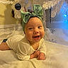 Elliana is registered to the contest to win money with this photo: baby, child, smile, green_bow, headband, white_blanket, christmas_tree, ornaments, holiday, decorations, happy, indoors, portrait, infant, cute, festive, soft_texture, twinkling_lights, lying_down, joyful