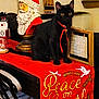 Fia is registered to the contest to win money with this photo: black_cat, cat, christmas, santa_statue, red_table_runner, holiday_decor, ornament, ribbon, bell, wooden_frame, sign, indoor, table, fabric, sitting, black_fur, gold_lettering, decorative_dove, wide_eyes, festive