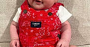 Maya is registered to the contest to win money with this photo: baby, infant, smiling, red_clothing, romper, sitting, couch, beige, texture, happy, cute, portrait, child, indoor, person, skin, face, small, young, apparel