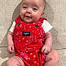 baby, infant, smiling, red_clothing, romper, sitting, couch, beige, texture, happy, cute, portrait, child, indoor, person, skin, face, small, young, apparel