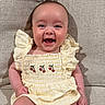 baby, child, smiling, yellow_outfit, ruffles, cherry_embroidery, couch, beige, textured_fabric, happy, sitting, indoors, cute, infant, portrait, young_child, human, skin, face, cheerful