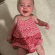 Maya is registered to the contest to win money with this photo: baby, child, smile, happy, dress, red, white, floral_pattern, infant, cute, lying_down, fabric, footwear, person, skin, indoors, texture, closeup, young, joy