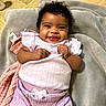 baby, smiling, curly_hair, blanket, purple_clothing, white_bow, infant, happy, lying_down, soft_texture, indoors, cute, child, toddler, skin, face, person, hands, closeup, cozy