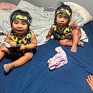 Gema And Bizus is registered to the contest to win money with this photo: toddler, child, bed, blanket, headband, sunflower, clothing, indoor, blue_sheet, gray_blanket, remote_control, shoes, plastic_shoes, happy, curious, sitting, person, face, hair, hand