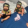 toddler, child, bed, blanket, headband, sunflower, clothing, indoor, blue_sheet, gray_blanket, remote_control, shoes, plastic_shoes, happy, curious, sitting, person, face, hair, hand