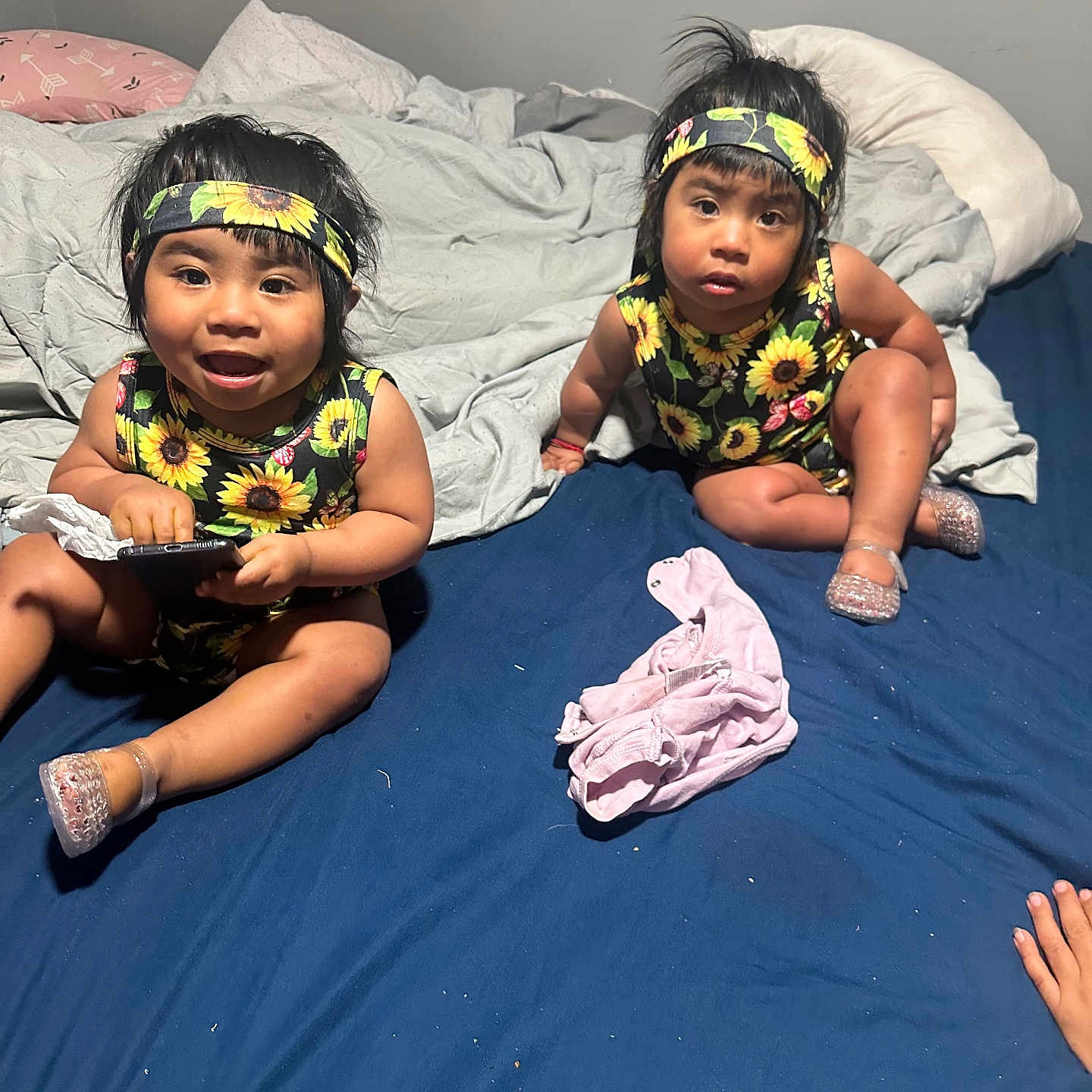 Gema And Bizus is registered to the contest to win money with this photo: toddler, child, bed, blanket, headband, sunflower, clothing, indoor, blue_sheet, gray_blanket, remote_control, shoes, plastic_shoes, happy, curious, sitting, person, face, hair, hand