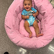 Malakaii is registered to the contest to win money with this photo: baby, smiling, blue_onesie, pink_chair, cushion, floor, tile_floor, indoor, child, happy, person, seat, plush, clothing, infant, cute, baby_face, furniture, relaxing, young_child