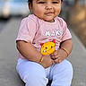 child, toddler, portrait, sitting, outdoor, pavement, smiling, face, hair, pink_shirt, white_pants, blue_socks, orange_trim, toy, plush_toy, bracelet, daylight, blurred_background, street, cute