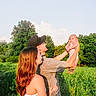 baby, casual_clothing, clouds, family, field, grass, greenery, happy, hat, holding, lifting, man, nature, outdoor, portrait, sky, summer, sunlight, tattoo, woman