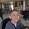 child, boy, smile, haircut, salon, barbershop, steering_wheel, chair, interior, tiles, person, mirror, cabinet, hair_washing_station, clothing, floor, door, lighting, happy, casual