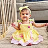 baby, barefoot, bow, bracelet, brown_eyes, child, couch, crib, curly_hair, flower_pattern, headband, indoors, nursery, pink_dress_details, portrait, rug, sitting, smiling, toddler, yellow_dress