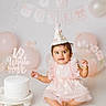 baby, child, birthday, birthday_hat, cake, pink_dress, balloons, party, celebration, portrait, smiling, sitting, infant, pastel, indoor, studio, decorations, white_cake, floral_details, happy