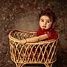 baby, child, portrait, sitting, wicker_bassinet, basket, red_dress, headband, bow, curly_hair, wide_eyes, facial_expression, indoor, studio, vintage, furniture, rattan, warm_lighting, textile_background, photography