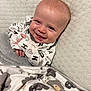 Eastyn is registered to the contest to win money with this photo: baby, smile, blanket, onesie, infant, face, cushion, happy, child, portrait, cozy, cute, lying_down, indoor, textile, soft, skin, head, person, toddler