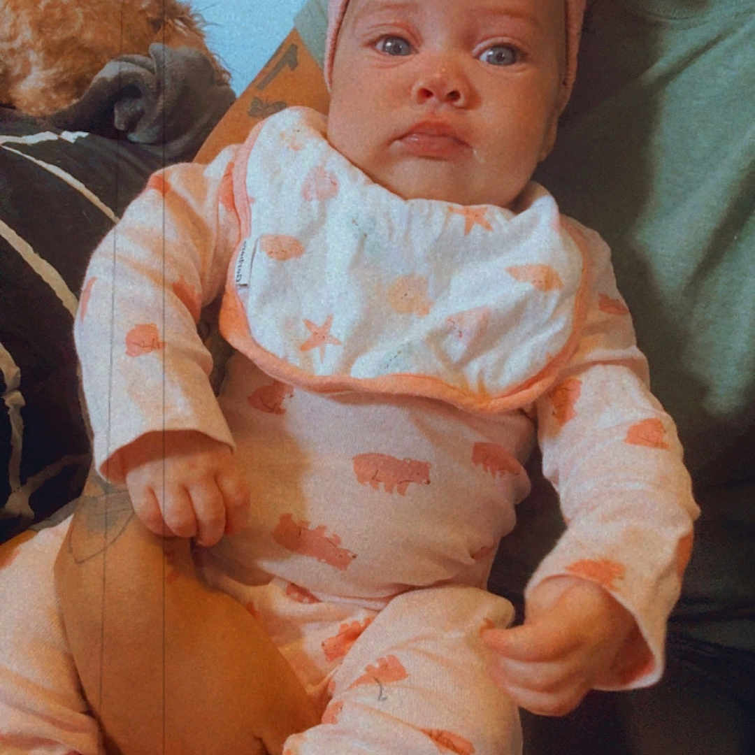 Jolie is registered to the contest to win money with this photo: baby, infant, child, portrait, sitting, close_up, wide_eyes, headband, bow, onesie, pink_clothing, patterned_clothing, sock, adult, lap, dog, pet, indoors, couch, family
