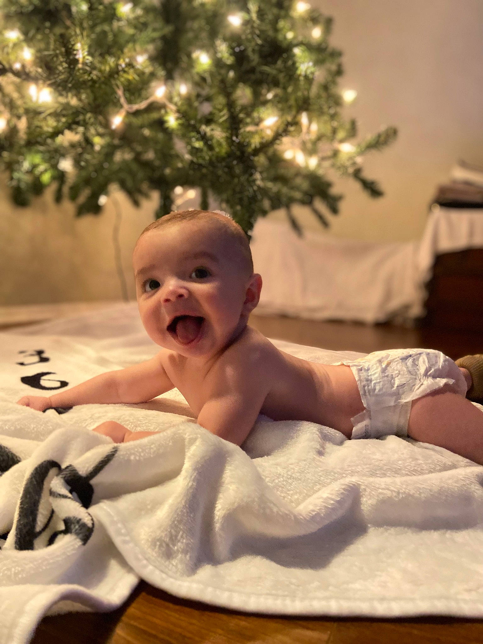 Addison is registered to the contest to win money with this photo: baby, baby_toddler_clothing, child, christmas_tree, comfort, event, finger, flooring, foot, fun, happy, linens, ornament, person, plant, room, sitting, skin, smile, toddler